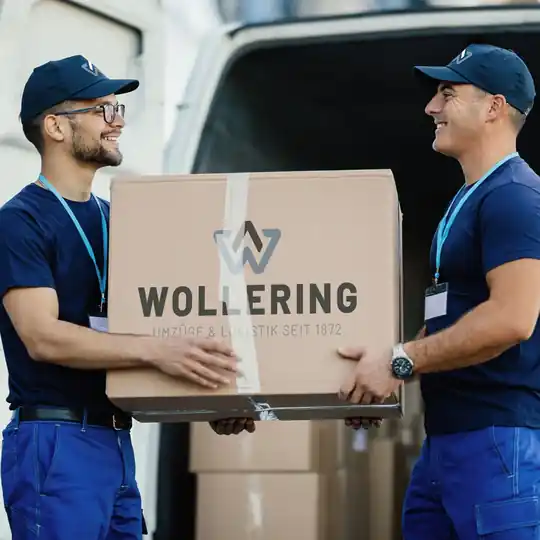Wollering-Umzugsteam trägt gemeinsam einen Karton mit Firmenlogo zum Umzugswagen